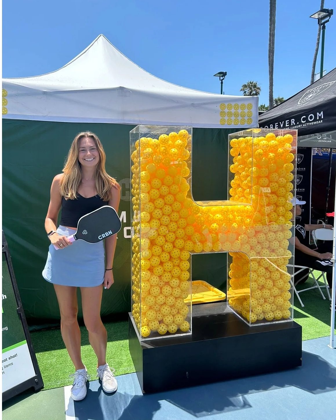 Hannah Ward at a tournament with her paddle, next to a giant letter H filled with pickleballs